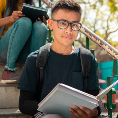 Outdoor image of Asian teenager, student sitting on stairs and doing study in the college campus day time. He is looking at the camera with a confident smile.